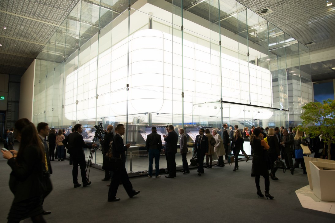 Attendees gather inside a modern glass-walled exhibition booth with illuminated curved panels at Baselworld.