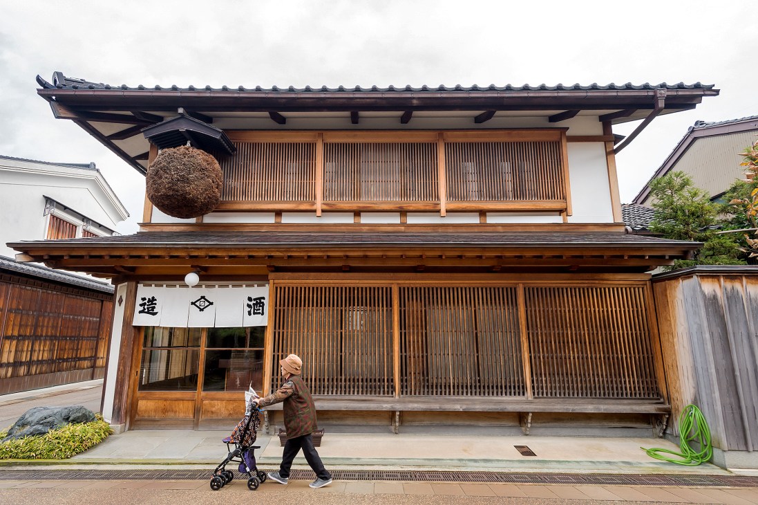 An elderly woman with a walker stands outside a traditional Japanese wooden building with a thatched roof decoration.