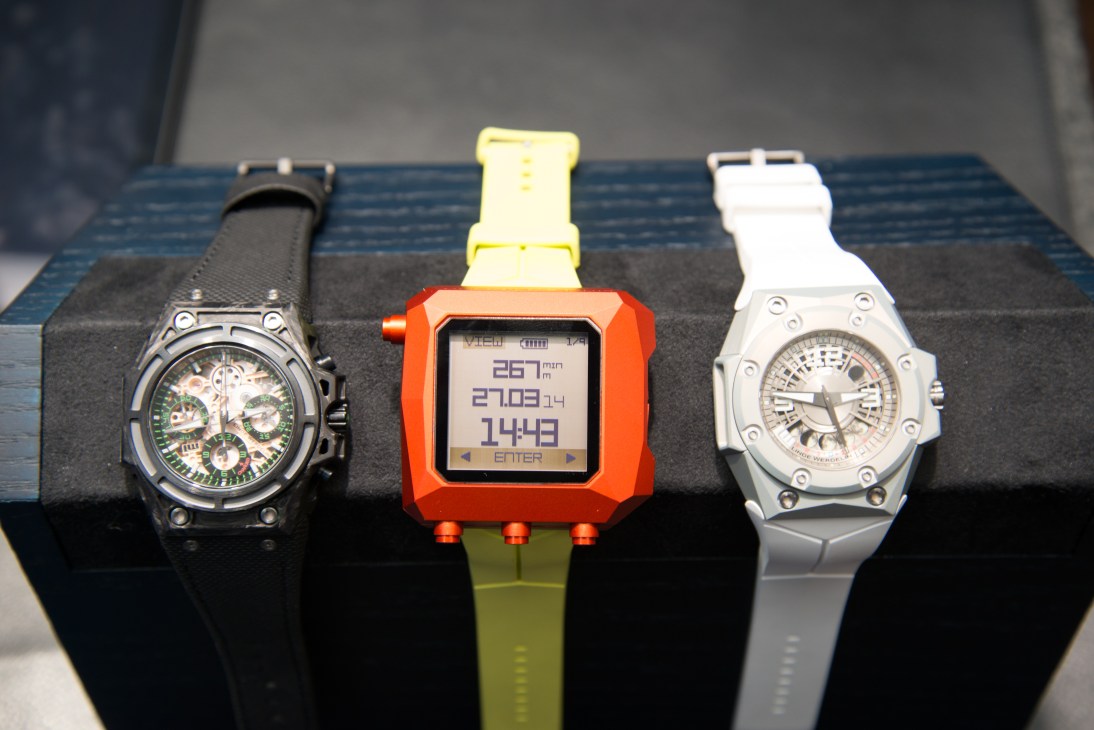 Three Linde Werdelin watches displayed on a stand: a black chronograph, an orange digital sports watch, and a white dial timepiece.