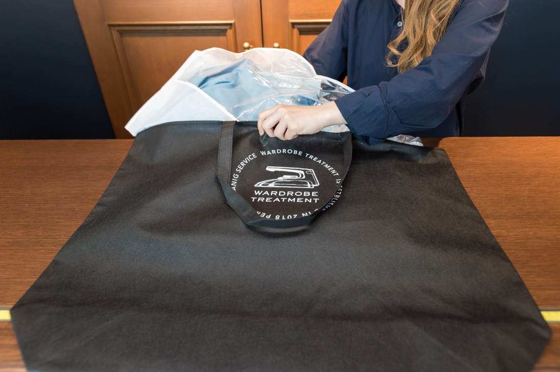 A staff member at Wardrobe Treatment places a cleaned garment in a branded black delivery bag on a wooden counter.