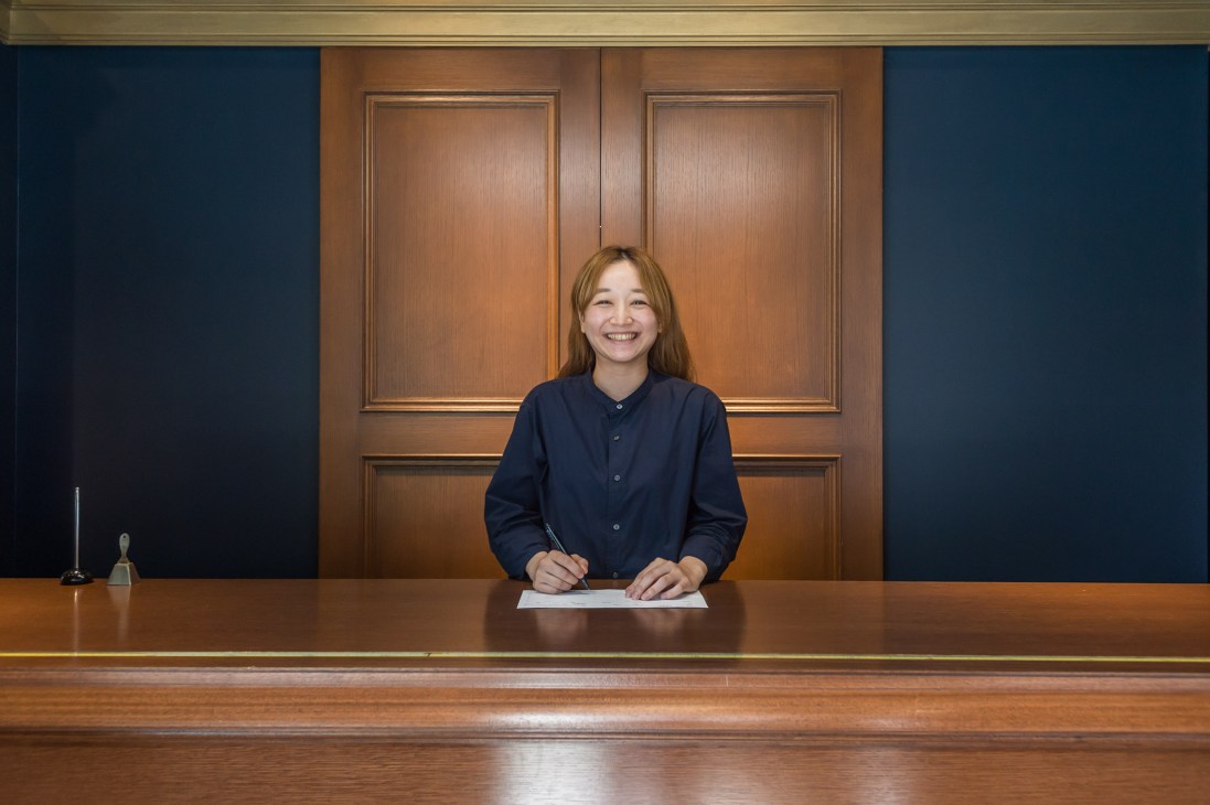 Woman in navy shirt stands at wooden counter in drycleaner shop with paneled wood wall behind.