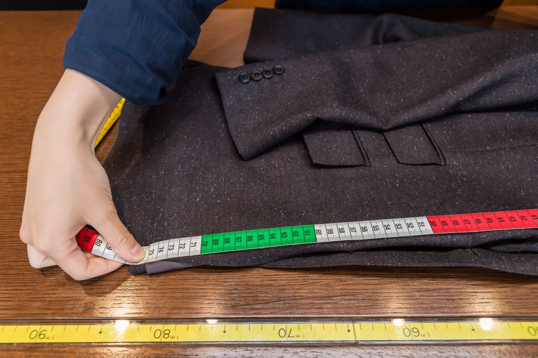 A person measures a dark suit jacket with a tape measure on a wooden table.