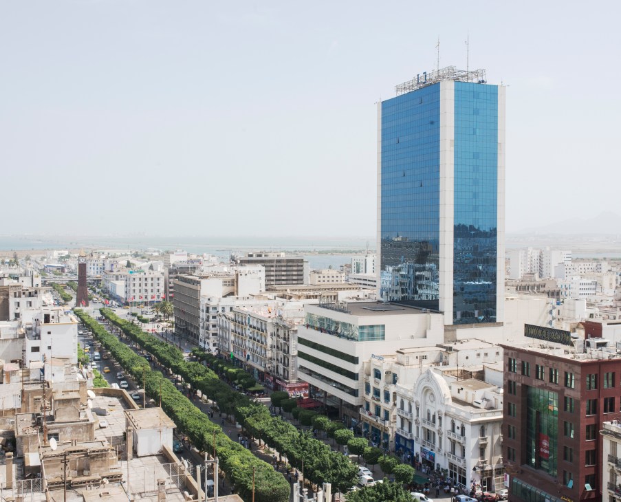 Avenue Bourguiba in Tunis with tree-lined street, modern blue glass tower, and historic buildings under hazy sky.