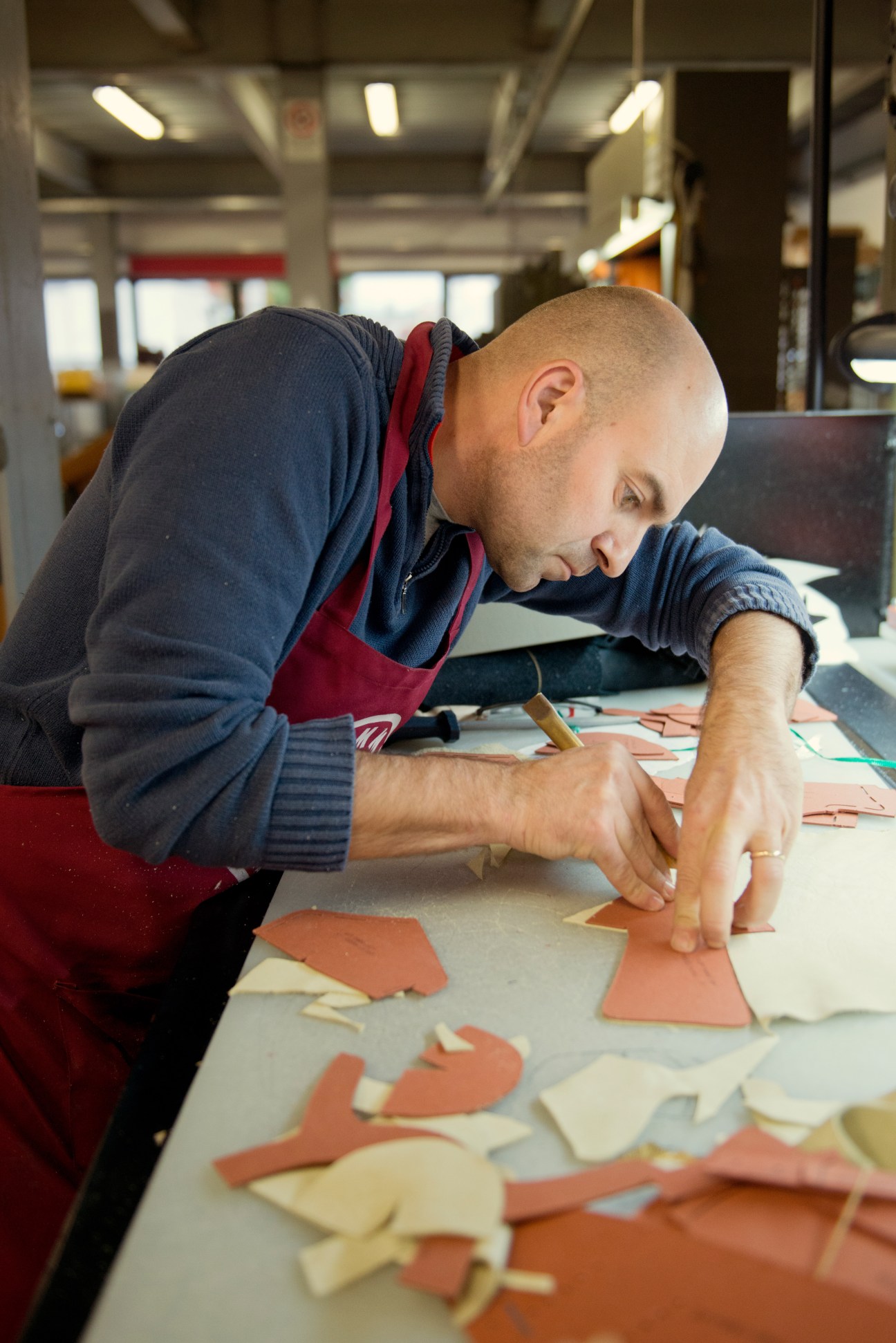 A worker in a red apron precision cuts leather pieces at a factory workbench.