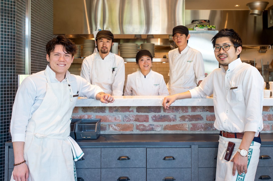 Five restaurant staff members in white uniforms standing behind a counter in a modern kitchen.
