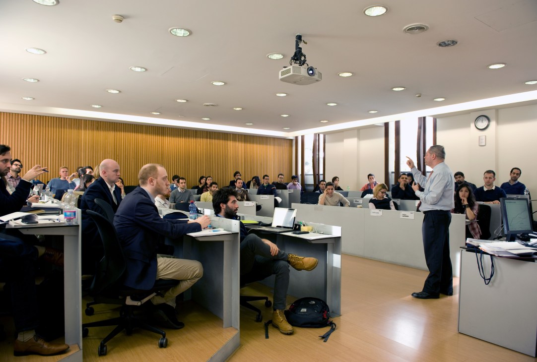 Professor teaching a management class to MBA students seated at individual desks in a modern classroom.
