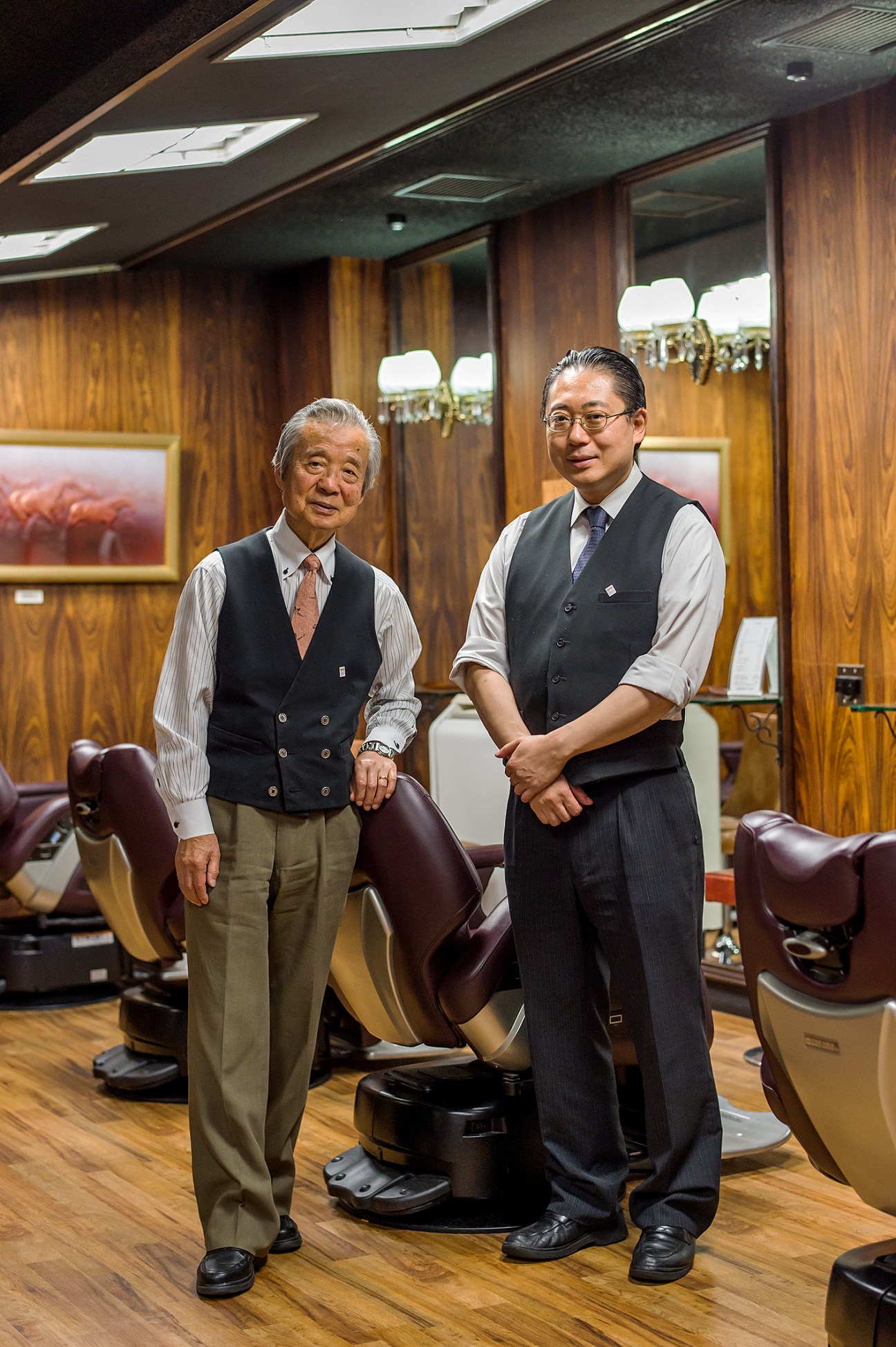 Two men in formal attire stand in a traditional barber shop with wooden paneling and vintage barber chairs.