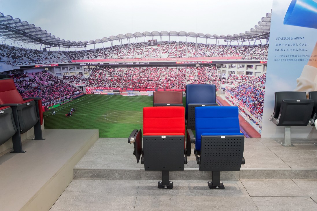 Stadium seating in red and blue colors displayed in front of a large soccer field with crowds in the background.