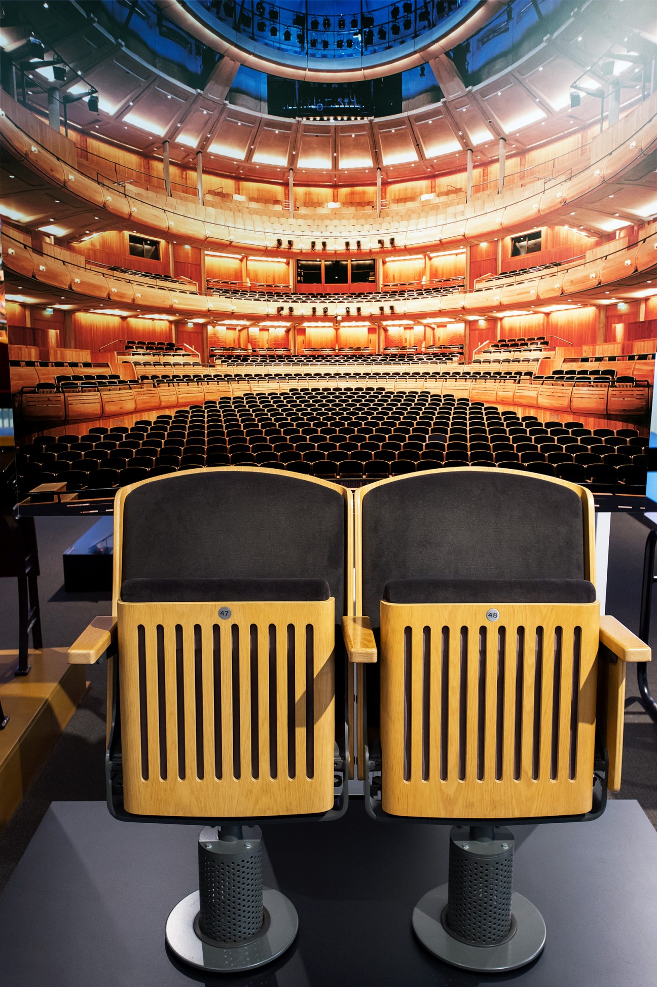Two wooden theatre seats with black upholstery in an empty auditorium with red-orange walls and multiple tiers of seating.