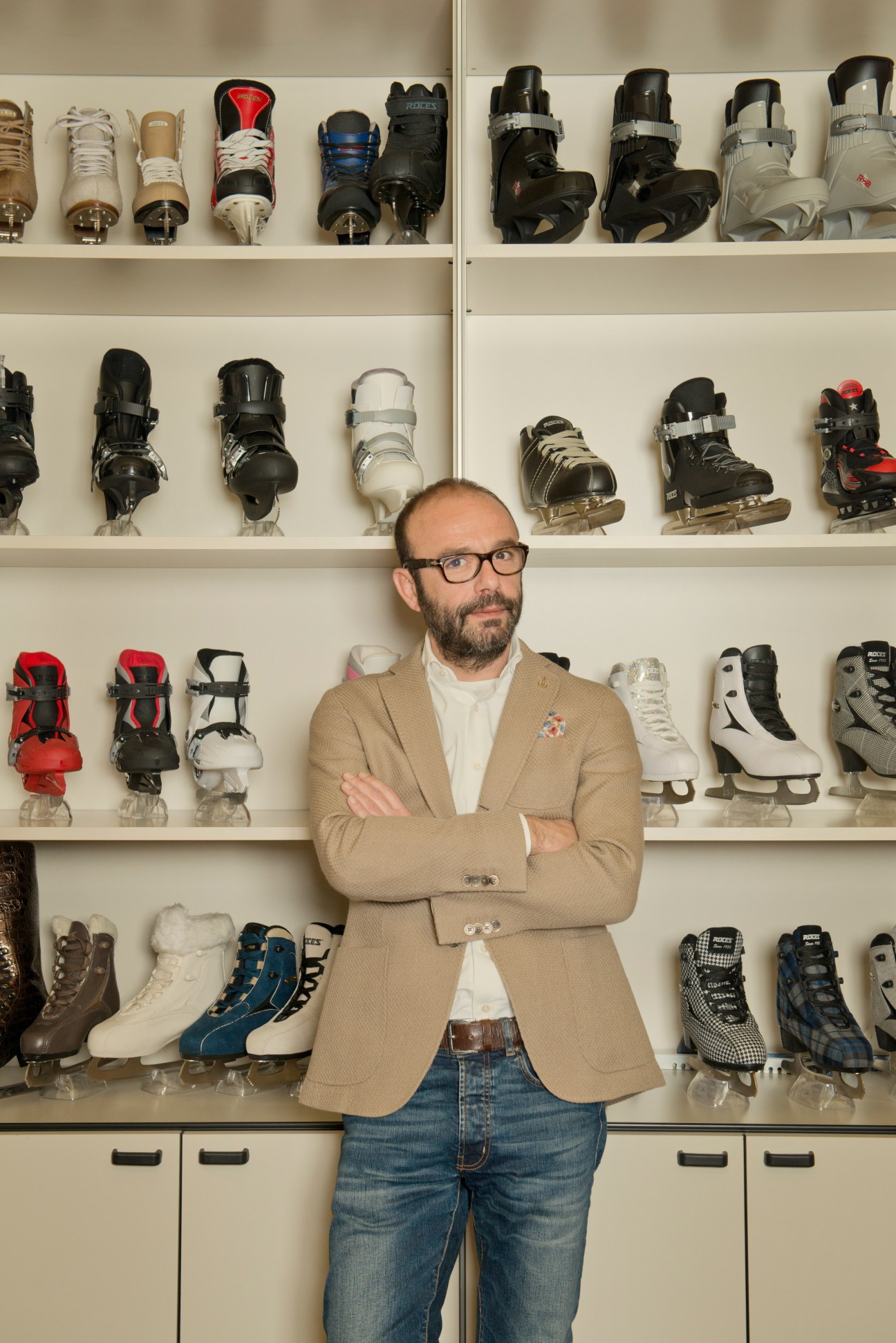 Man with glasses in tan blazer stands with arms crossed in front of wall of ice skates.