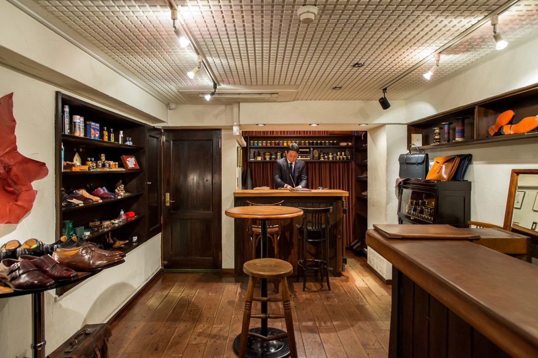 Interior of Brift H shoe-care salon in Tokyo with wooden counter, shelving, and a man in suit at desk.