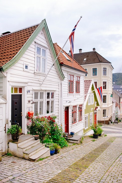 Colorful traditional wooden homes on a cobbled street in Bergen's Nordnes neighborhood with Norwegian flags displayed.