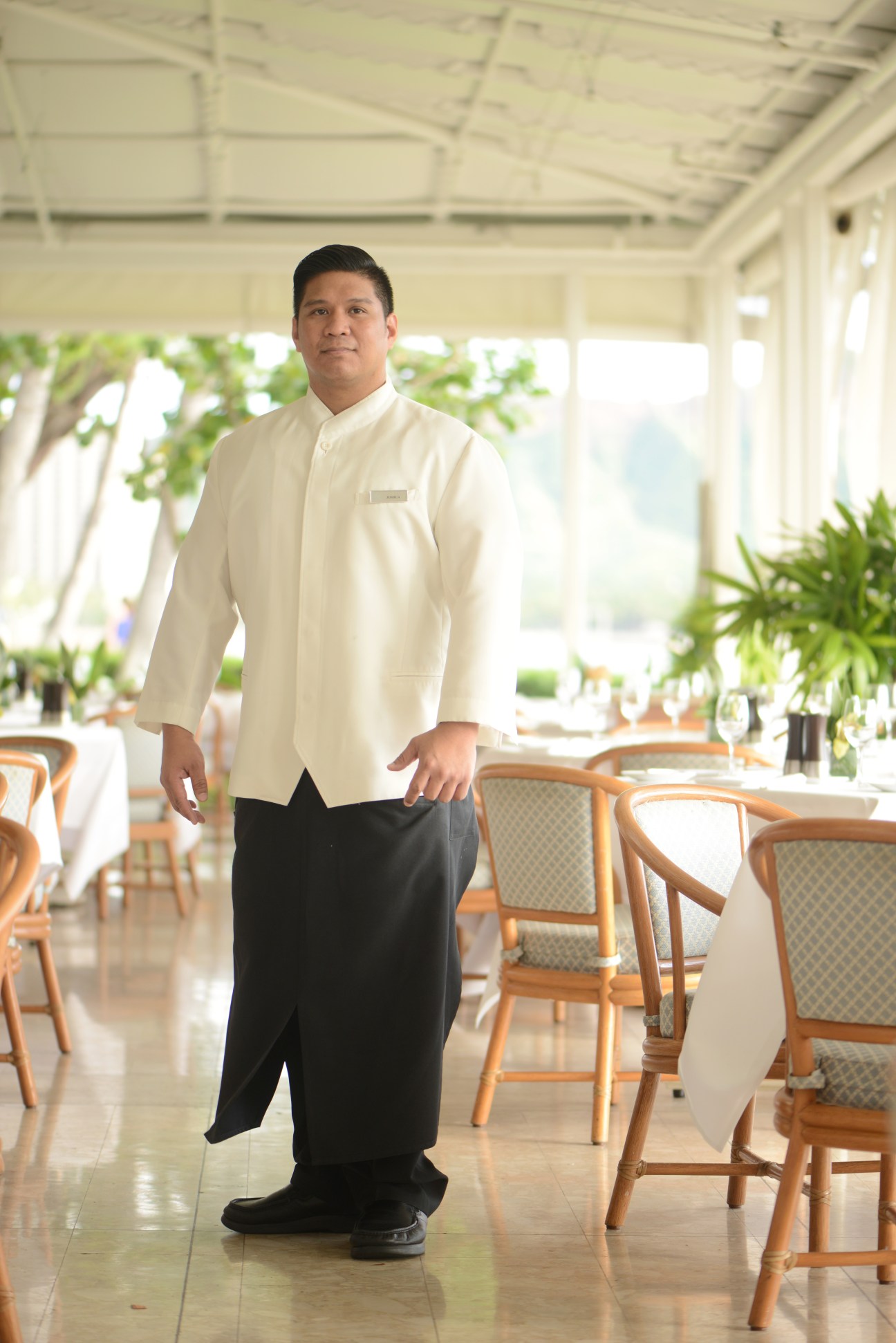 Man in cream chef's jacket and black trousers standing in bright dining room with wooden chairs.
