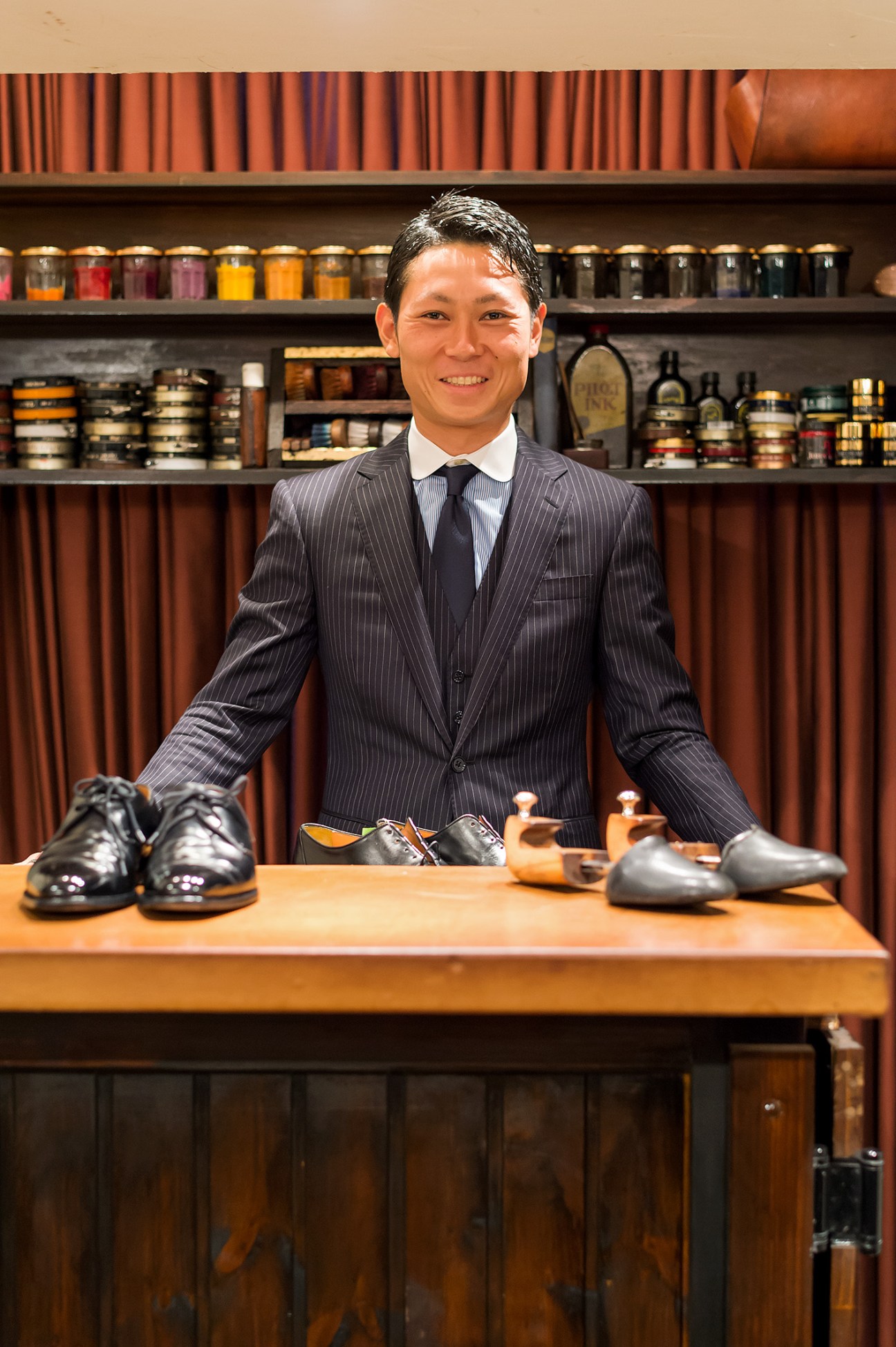 Man in pinstriped suit standing behind counter with polished shoes and shoe care tools displayed.