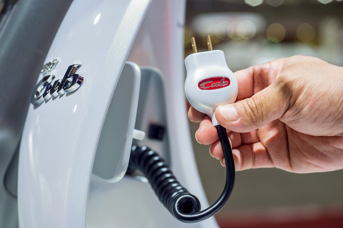 Hand holding a white charging plug with red Cub logo next to metallic-white Honda EV-Cub scooter.