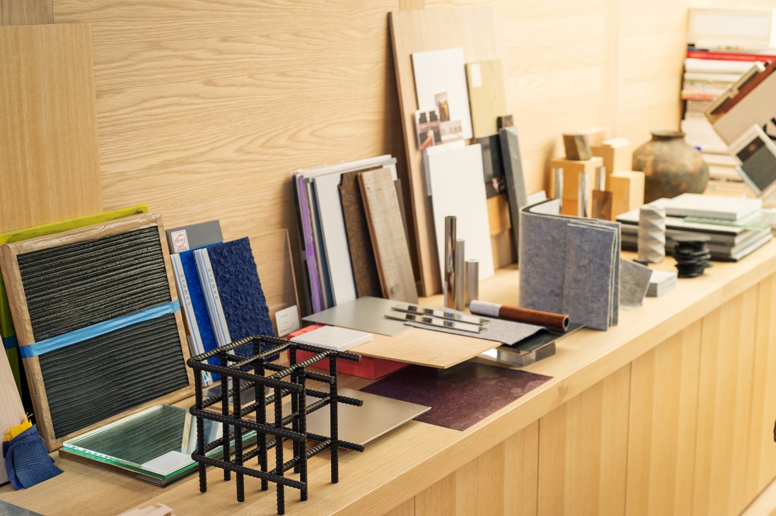 Design workspace with material samples, fabric swatches, and a miniature steel-rod-and-acrylic table model on a wooden desk.