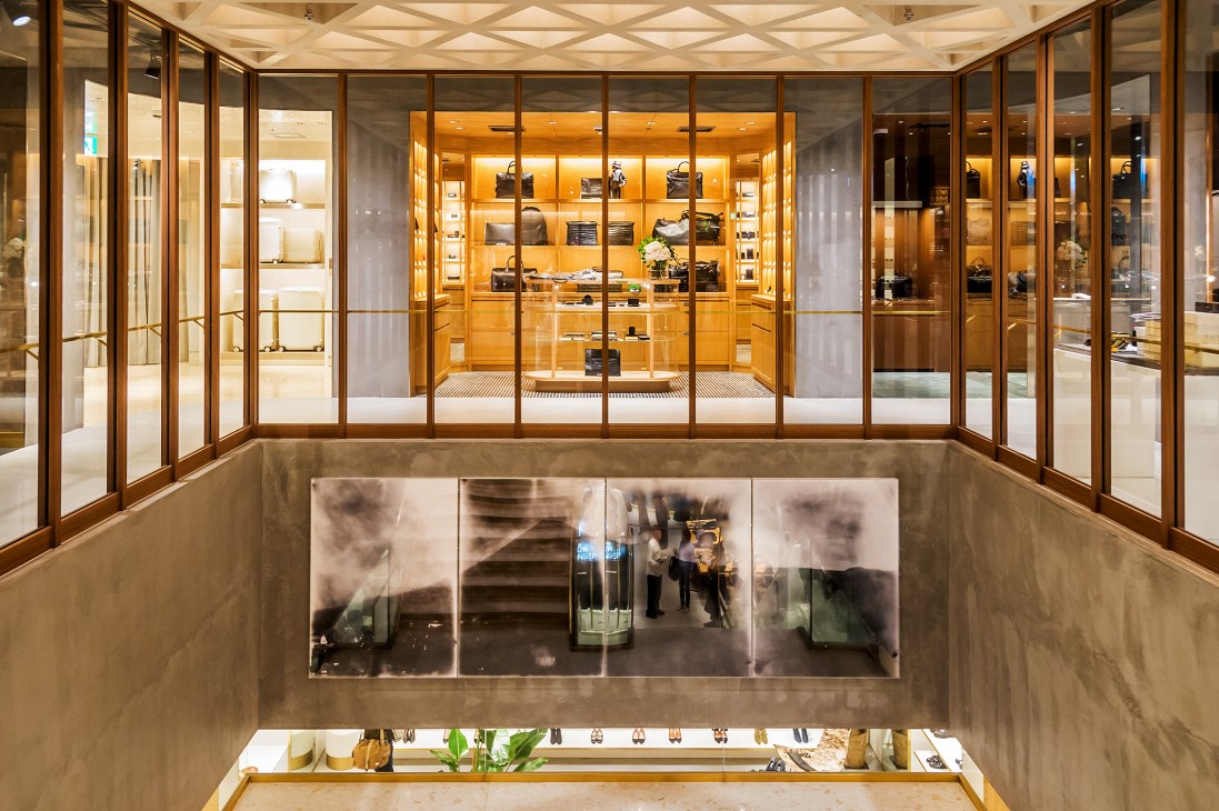 Multi-level retail store interior with glass-walled upper floor displaying luggage and leather goods