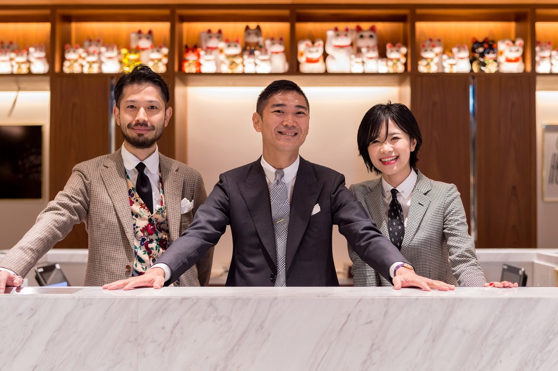 Three smiling staff members in suits standing behind marble counter with decorative cat figurines displayed on shelves