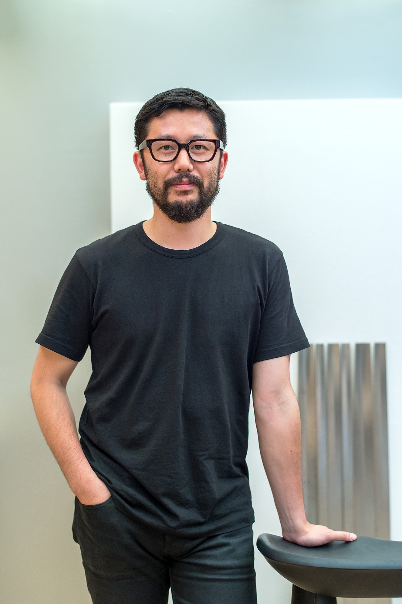 Portrait of Keisuke Nakamura, a man with dark hair and beard wearing glasses and a black t-shirt, standing in a minimalist interior space.