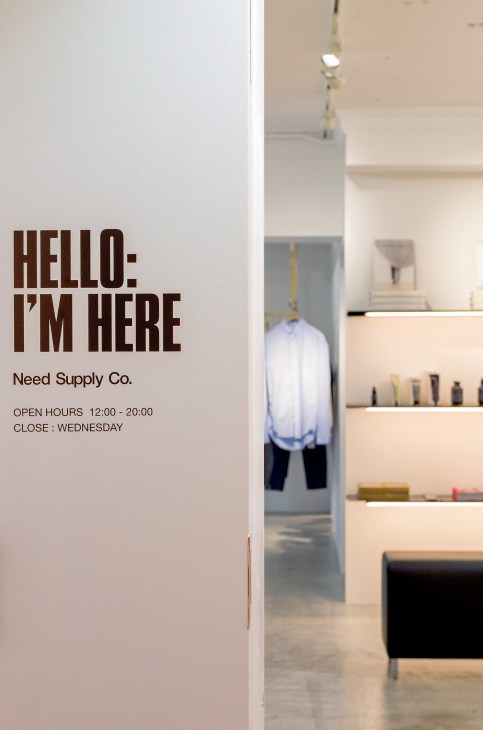 Need Supply Co storefront entrance with "HELLO- I'M HERE" sign and open hours posted on white wall.