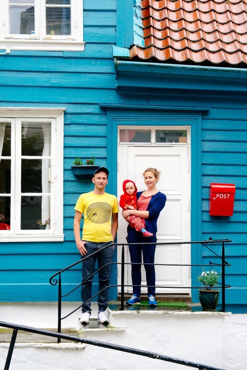 A man in a yellow shirt and woman holding a child in red stand outside a bright blue wooden house with a white door and red postbox.
