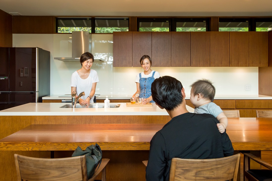 Two women stand behind a kitchen island counter while a man and child sit at the dining table in a modern wooden kitchen with white backsplash.