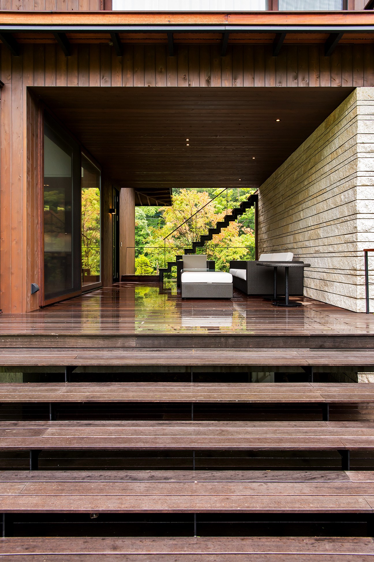 Modern wooden deck with black lounge furniture overlooking a forested landscape through large glass doors.