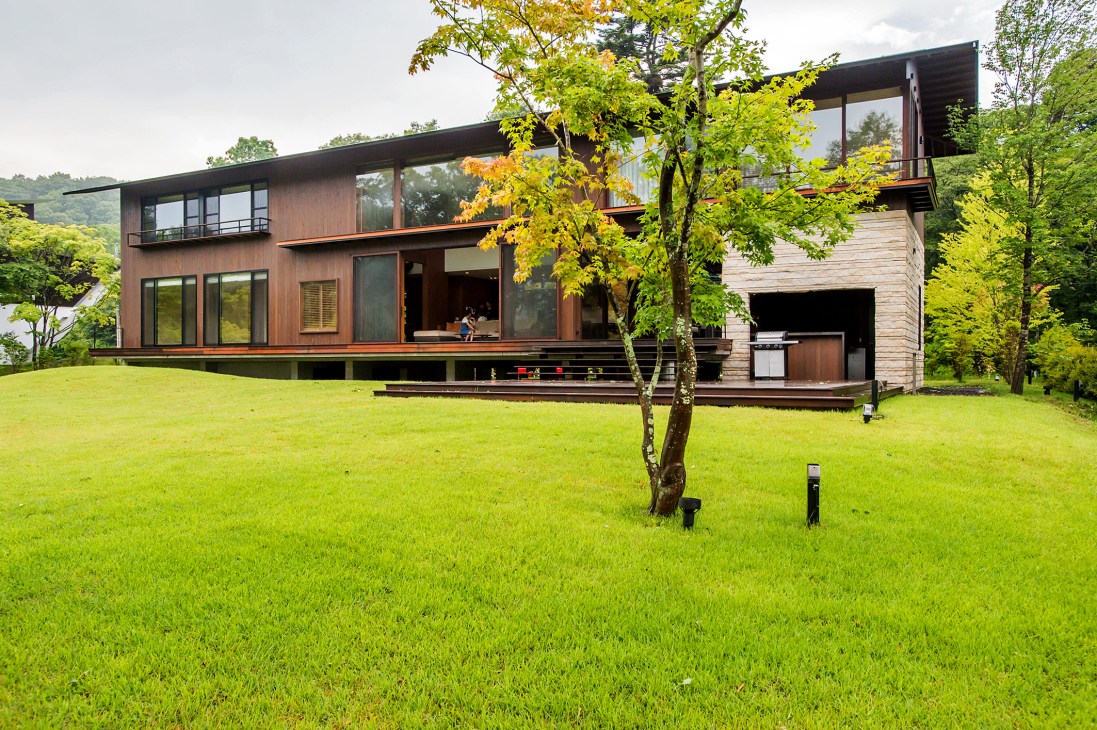Modern wooden house with large glass doors overlooking a manicured lawn and golf course beyond, surrounded by trees.