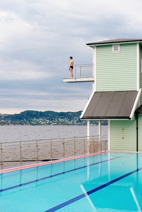 A swimmer stands on a diving platform at Nordnes Sjøbad, a heated saltwater bathing club with views over Bergen's fjord and hillside homes.