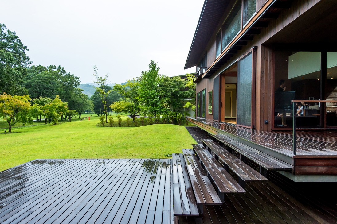 Modern wooden house with sliding glass doors opening onto a spacious deck overlooking a manicured lawn and forested golf course in Karuizawa.
