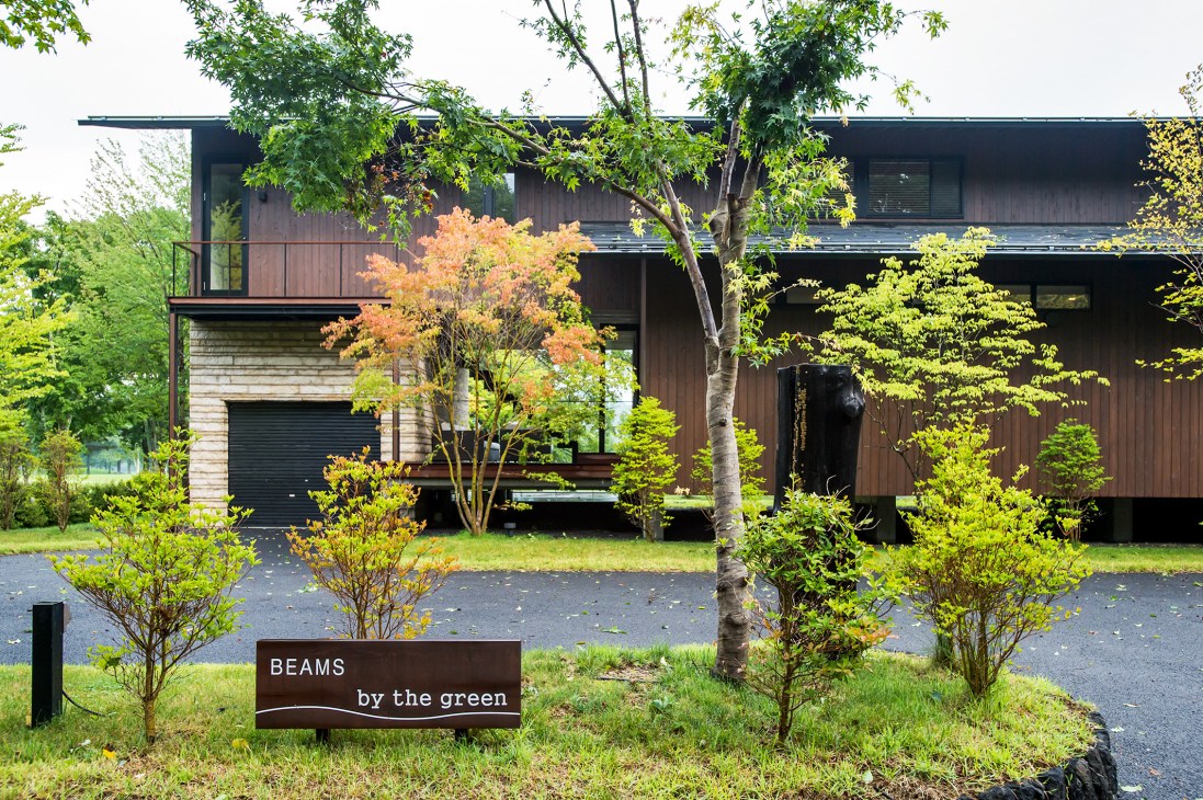 Dark wooden two-storey house with "Beams by the green" sign surrounded by trees and lawn.