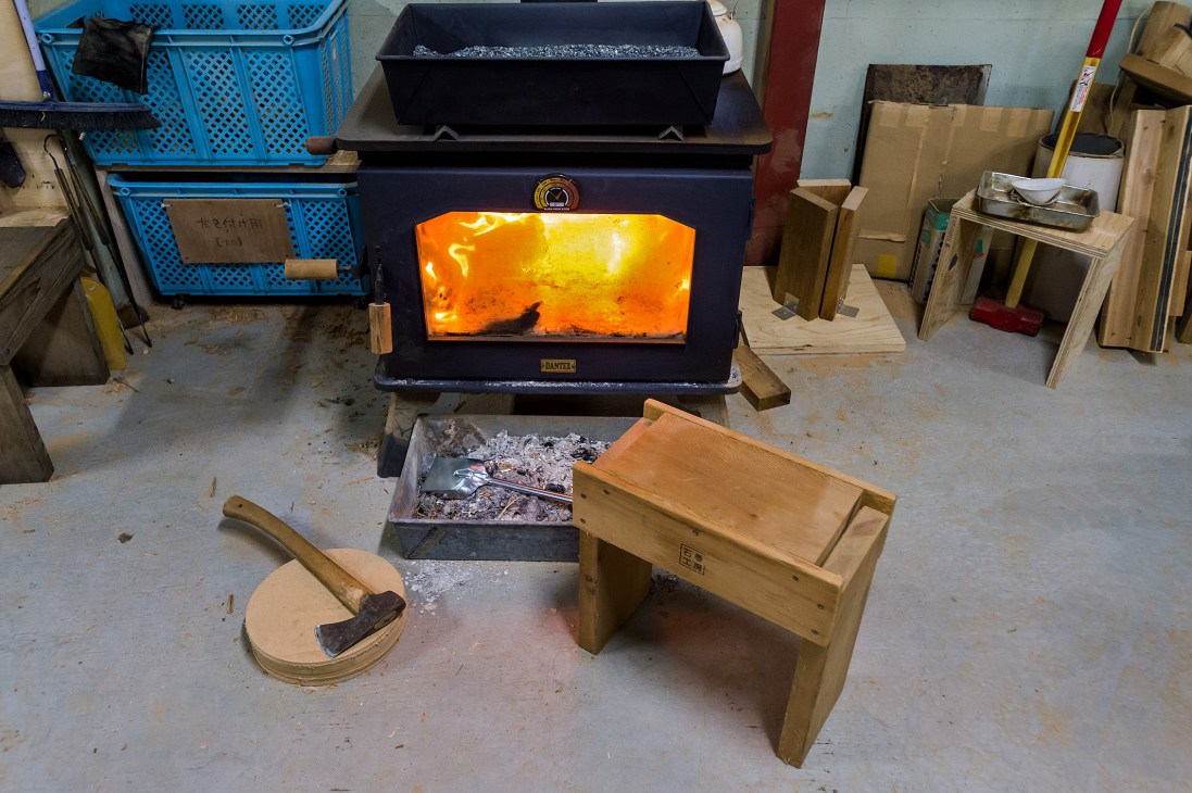 Wood-burning stove with glowing flames in Ishinomaki Laboratory workshop surrounded by wooden furniture and tools.