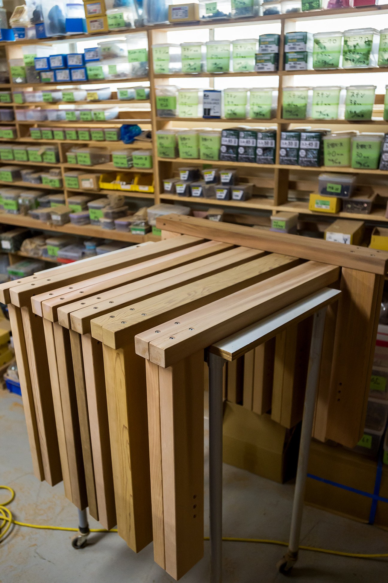 Wooden stools with slots stacked in rows inside a workshop with shelves of supplies in the background.