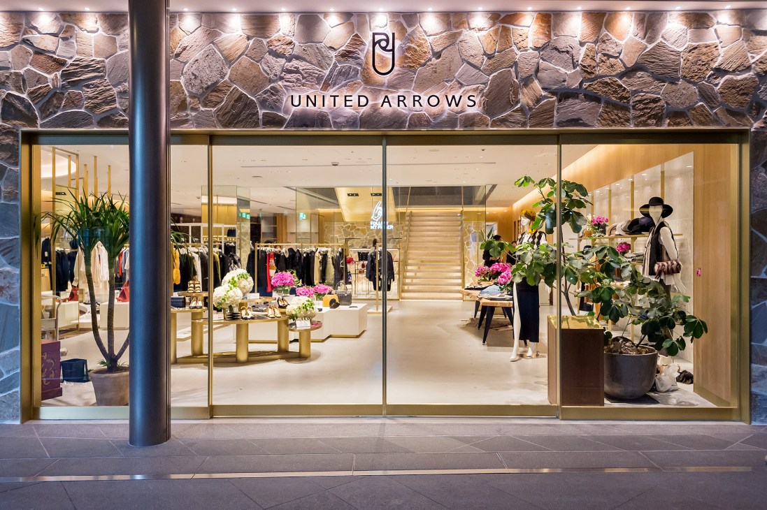 United Arrows storefront with stone facade, large windows displaying clothing racks and golden interior lighting