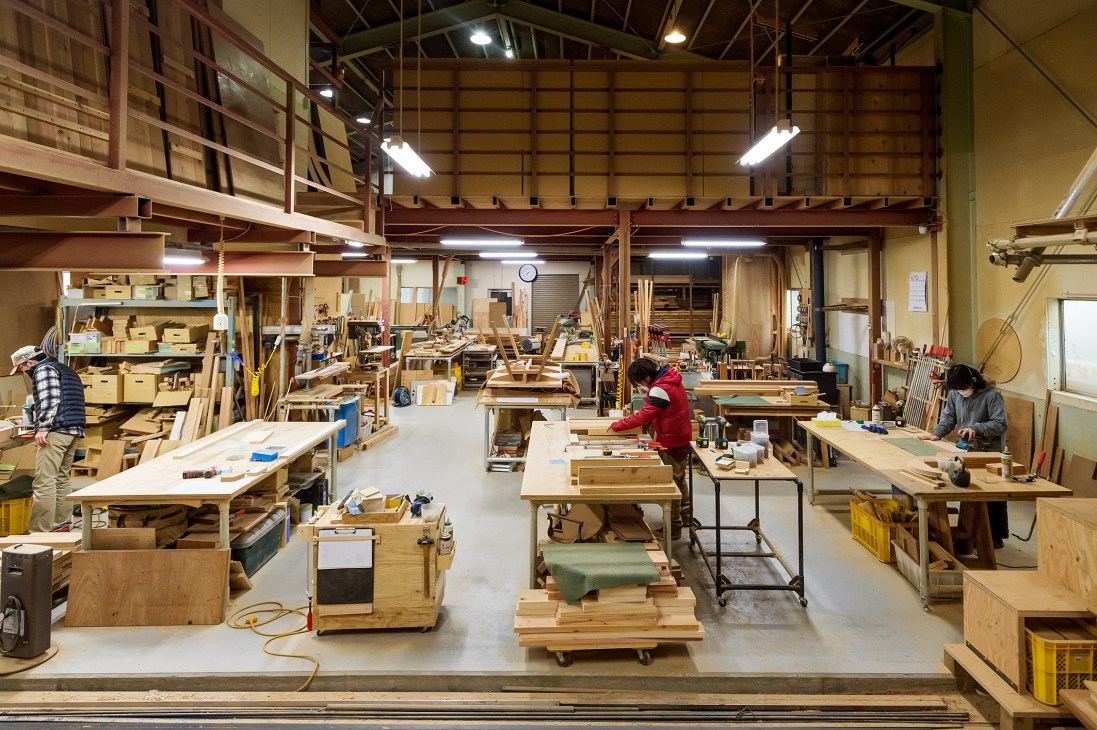 Interior of Ishinomaki Laboratory workshop with six craftspeople working at various wooden workstations among materials and tools.