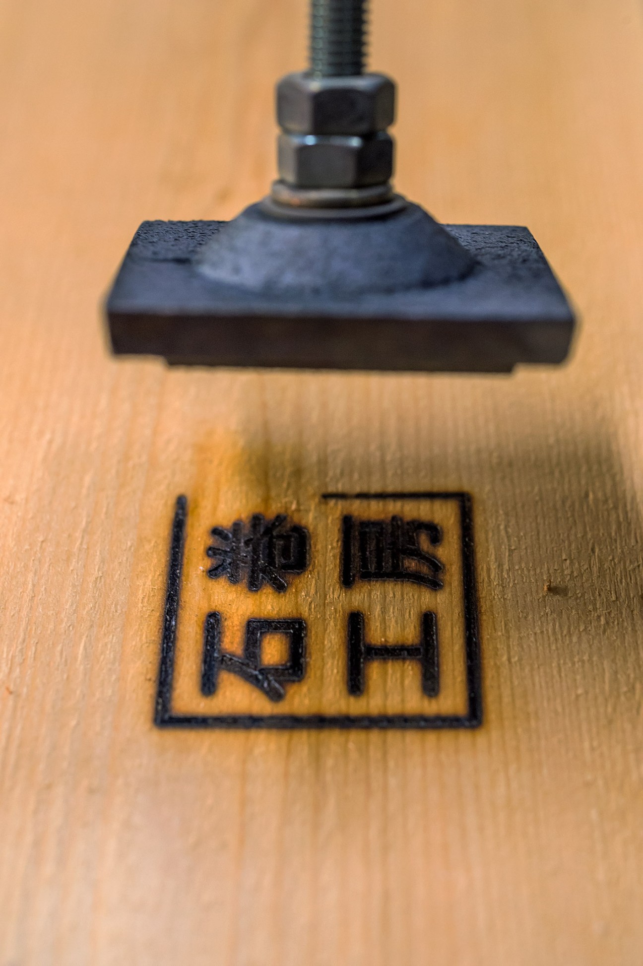 Close-up of a hot iron branding tool pressing Japanese characters onto light wood surface.