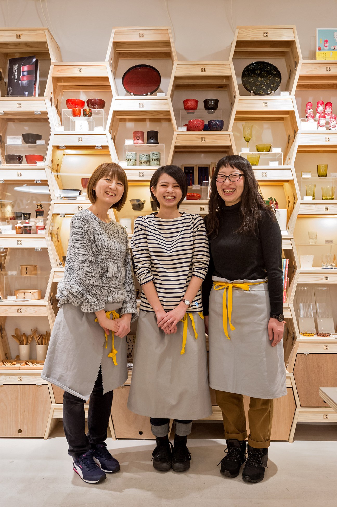 Three women wearing Ishinomaki Laboratory aprons stand smiling in front of wooden shelving display.