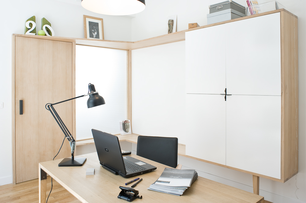 Modern office workspace with pale oak furniture, white panels, black desk lamp and laptop on wooden desk.