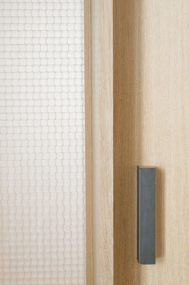 A rectangular grey block door handle on pale oak wood next to a translucent window panel.