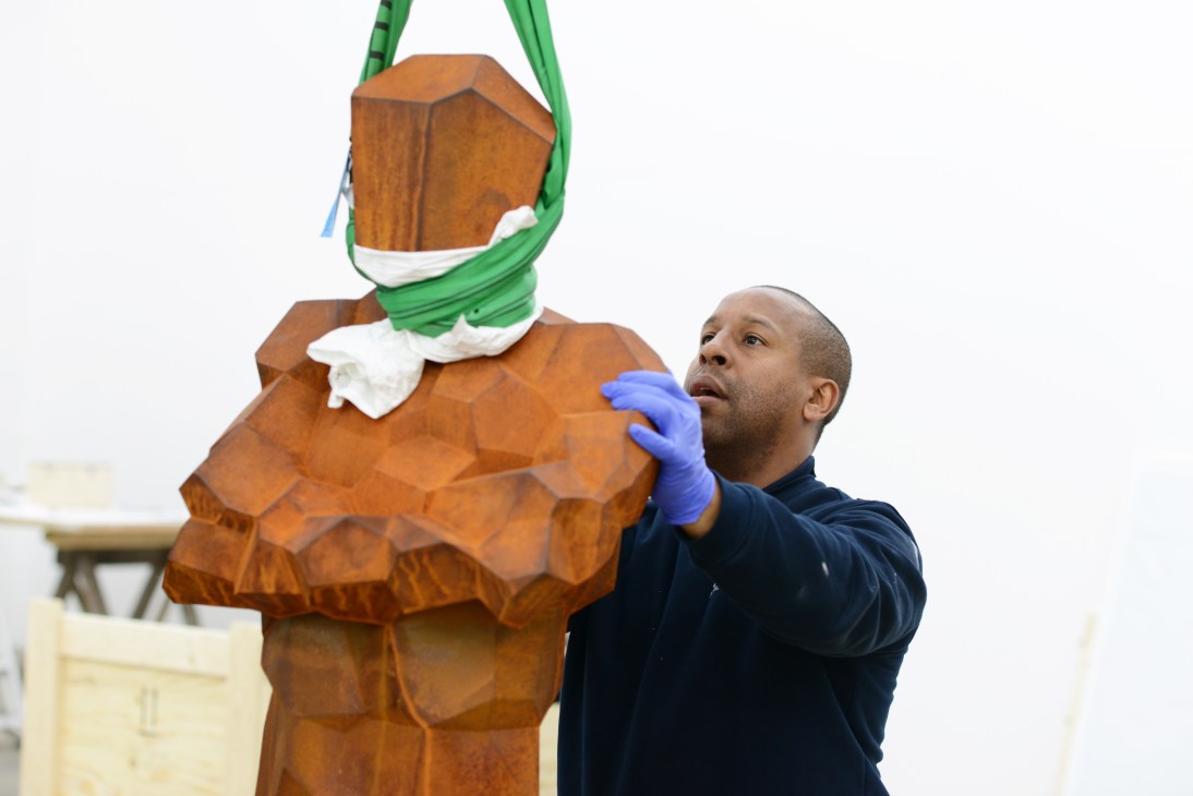 Art handler Danny Gayle in blue gloves carefully guides a suspended Antony Gormley iron sculpture during transport.