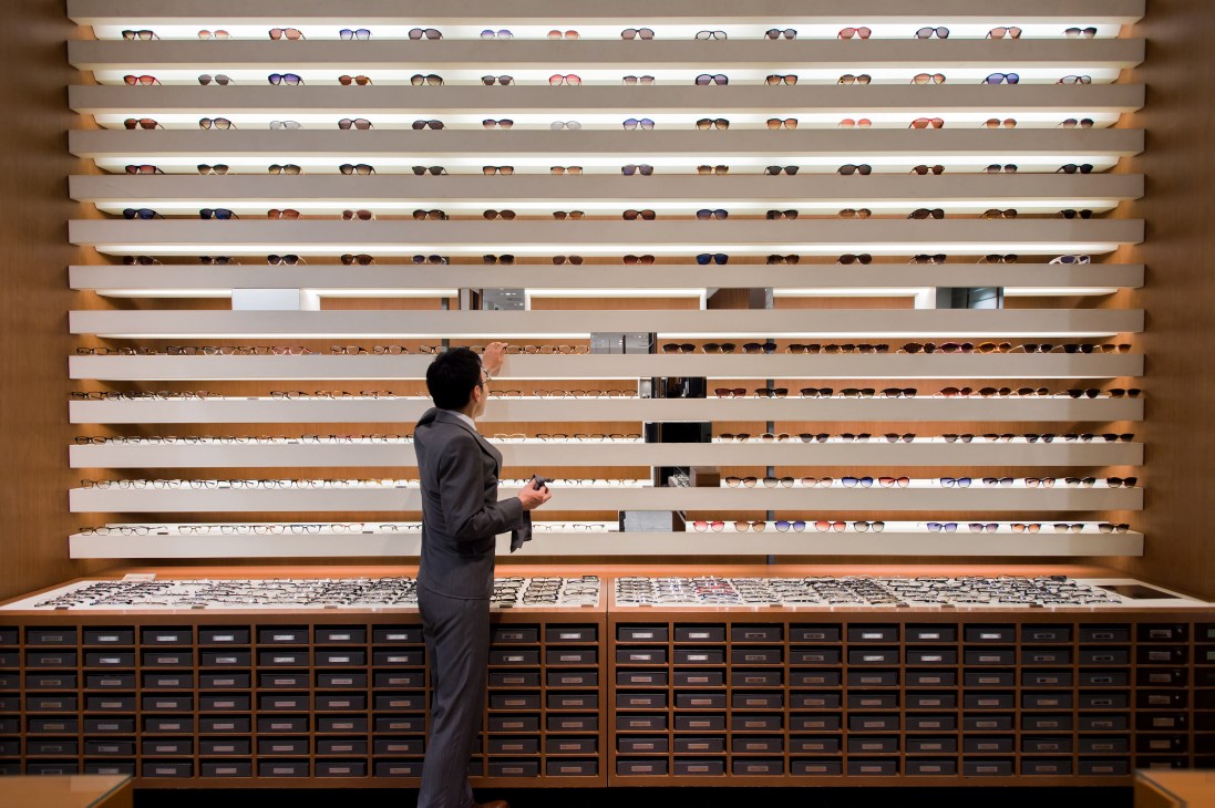 Staff member browsing organized eyewear display with horizontal shelves of sunglasses and frames at Isetan department store.
