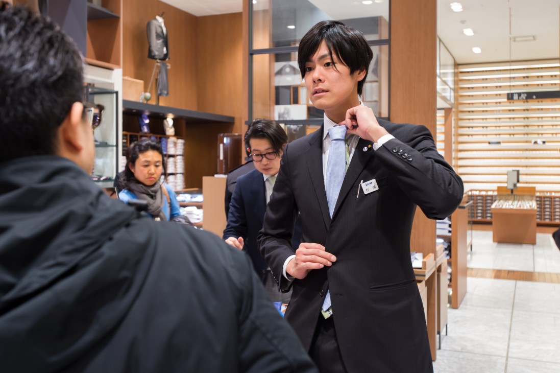 Male Isetan stylist in black suit and blue shirt assists customer in modern wood-paneled department store section.