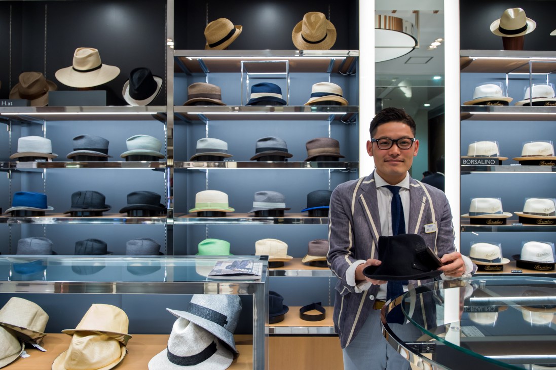 Male sales associate holding a hat at Isetan department store with shelves of various hats behind him.
