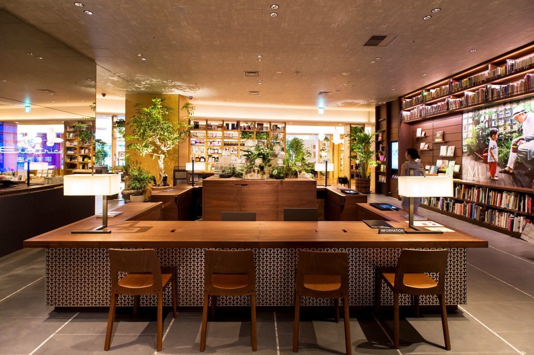 Interior of Tsutaya Electrics concierge desk with wooden counter, chairs, bookshelves, plants, and warm lighting.