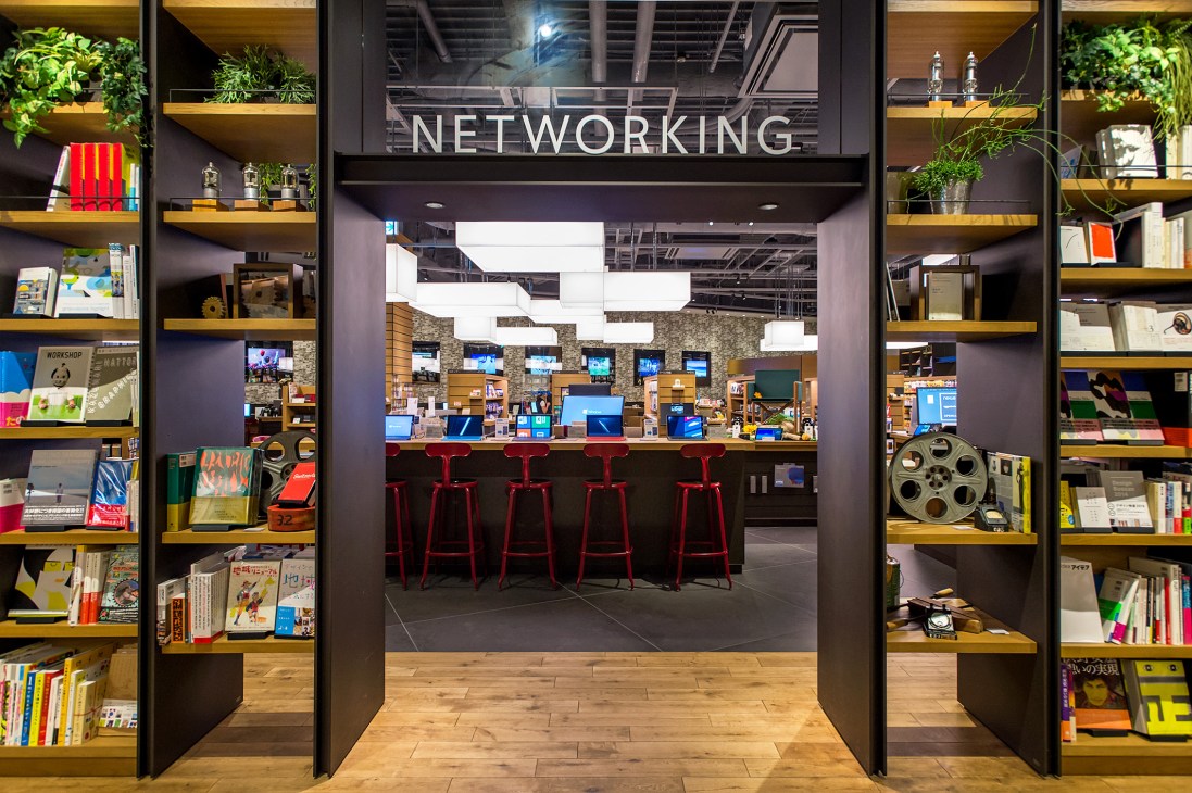 Tsutaya Electrics networking department with computers displayed at a service counter with red stools, surrounded by shelving with books and products.