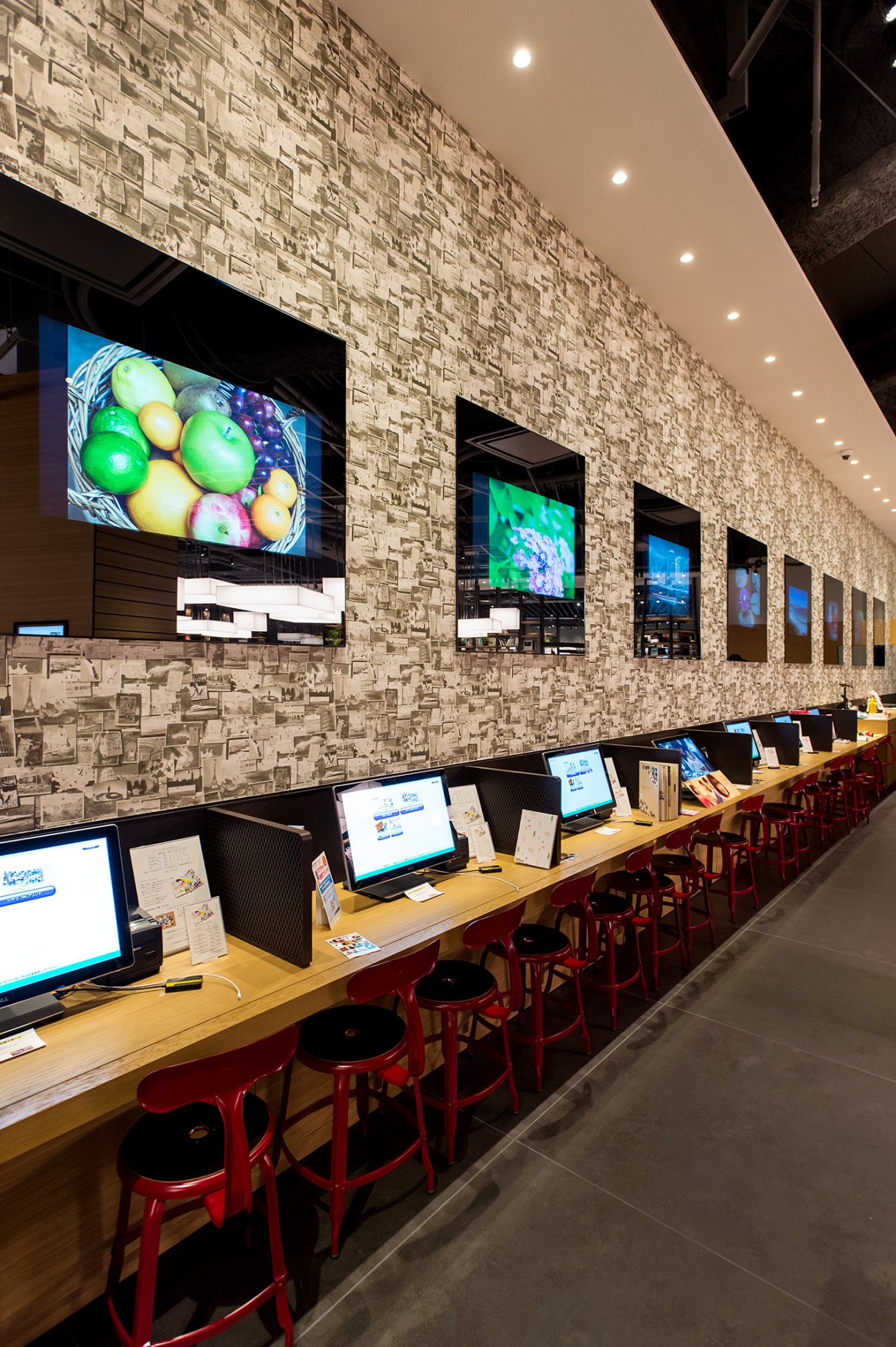 Photo-printing stations at Tsutaya Electrics with computers at wooden desks and red chairs, displaying colorful images on wall-mounted screens.