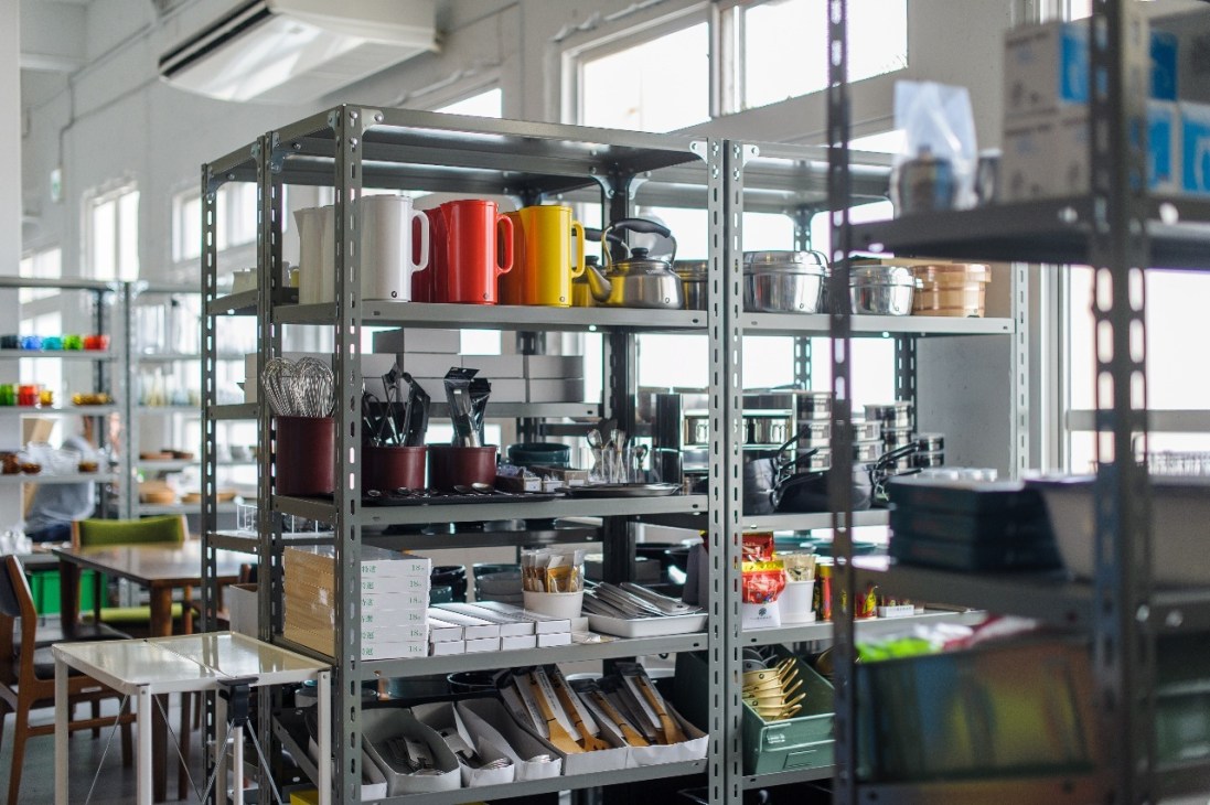 Industrial metal shelving unit displaying colorful kitchenware, including red and yellow mugs, cookware, and utensils in a bright, modern space.