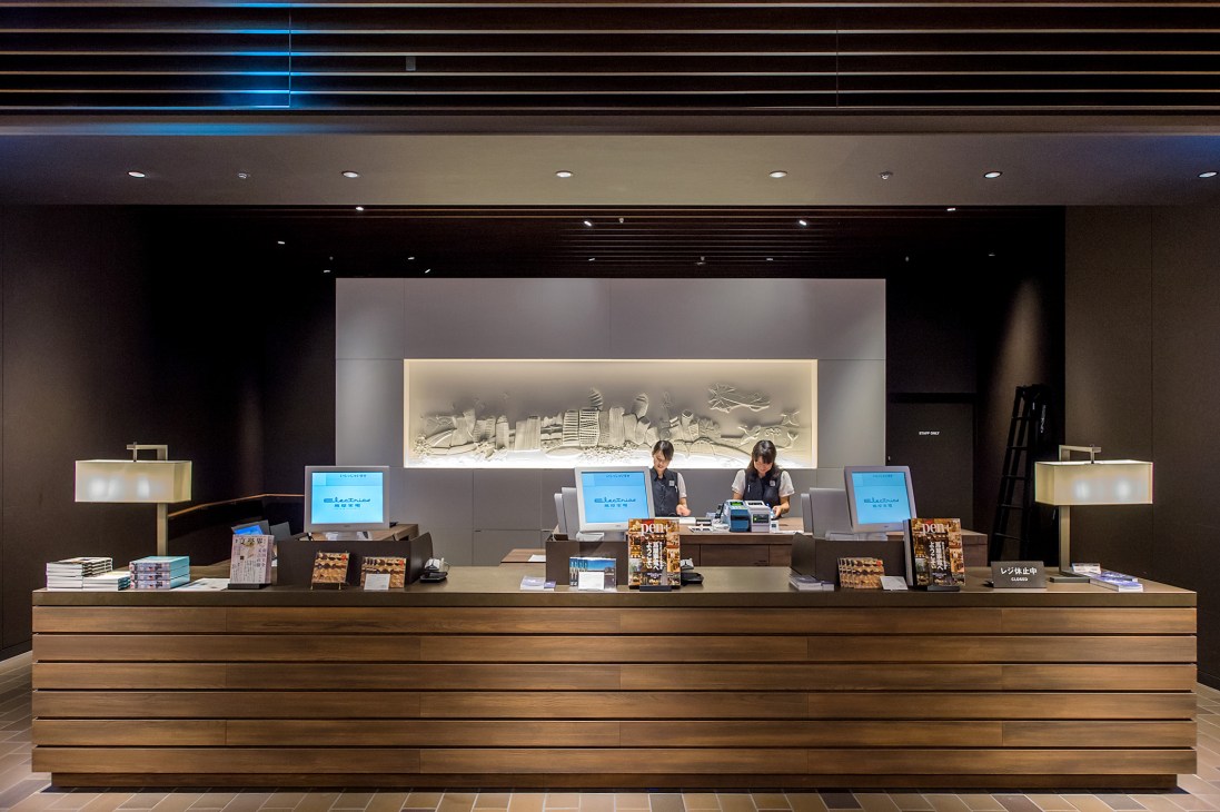 Tsutaya Electrics ground floor checkout desk with staff members, blue screens, and a beige-toned relief artwork of a cityscape on the back wall.