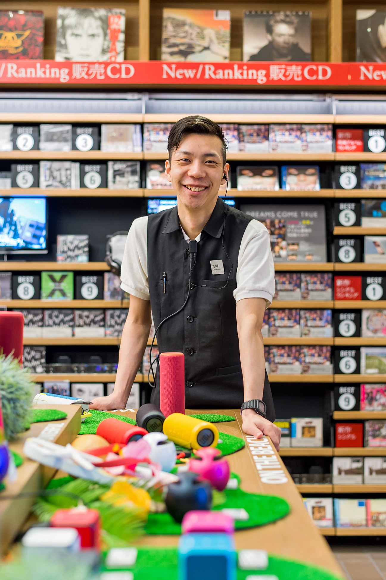 Smiling staff member at Tsutaya Electrics music section with colorful speakers and CDs displayed behind.
