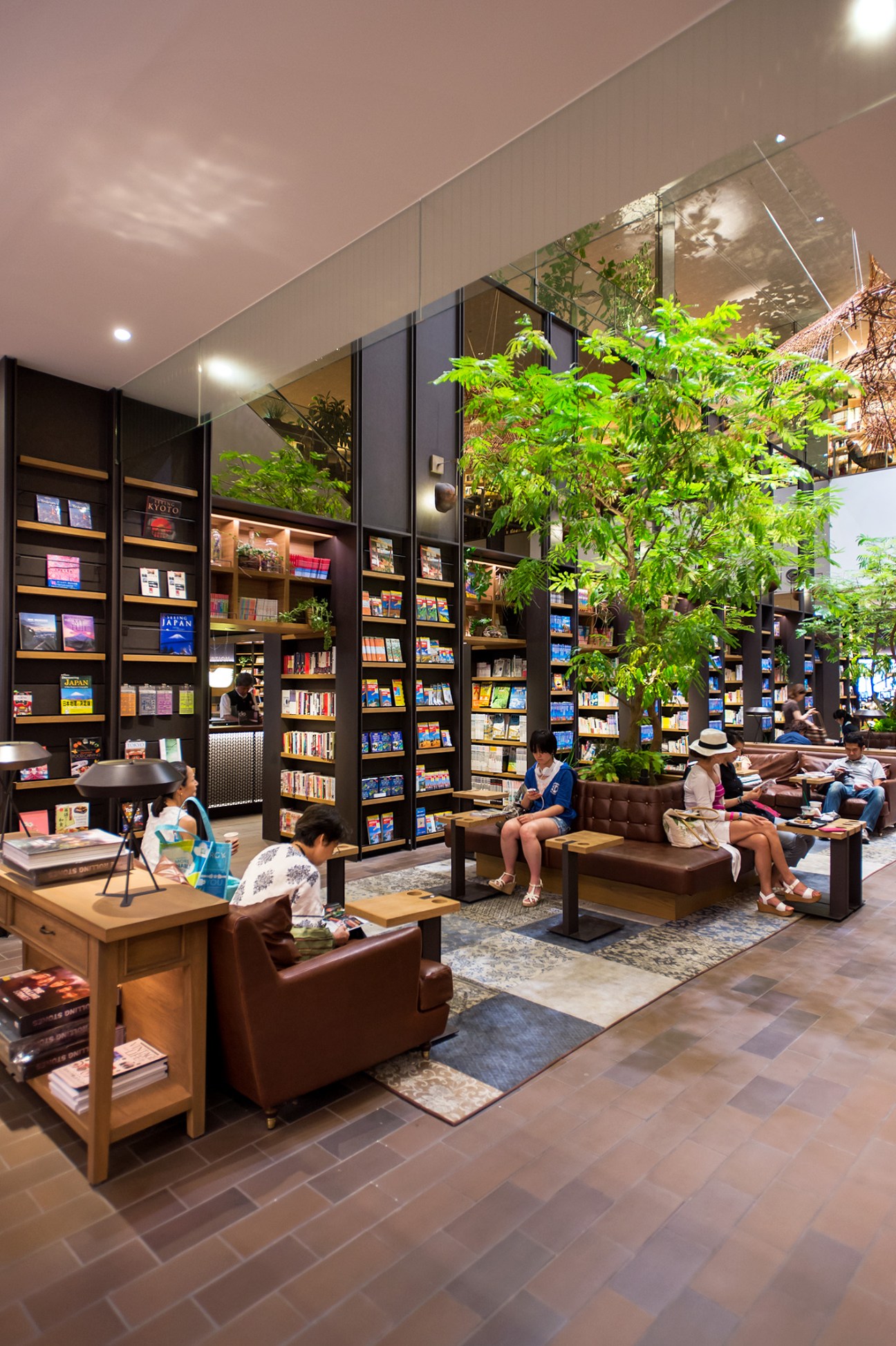 Modern bookstore interior with tall black shelving units filled with books, lush green plants, brown leather sofas, and customers browsing.